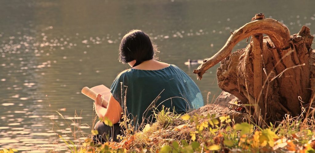A woman sits reading alone on the banks of a river with autumn leaves on the ground. Her back is to the camera.